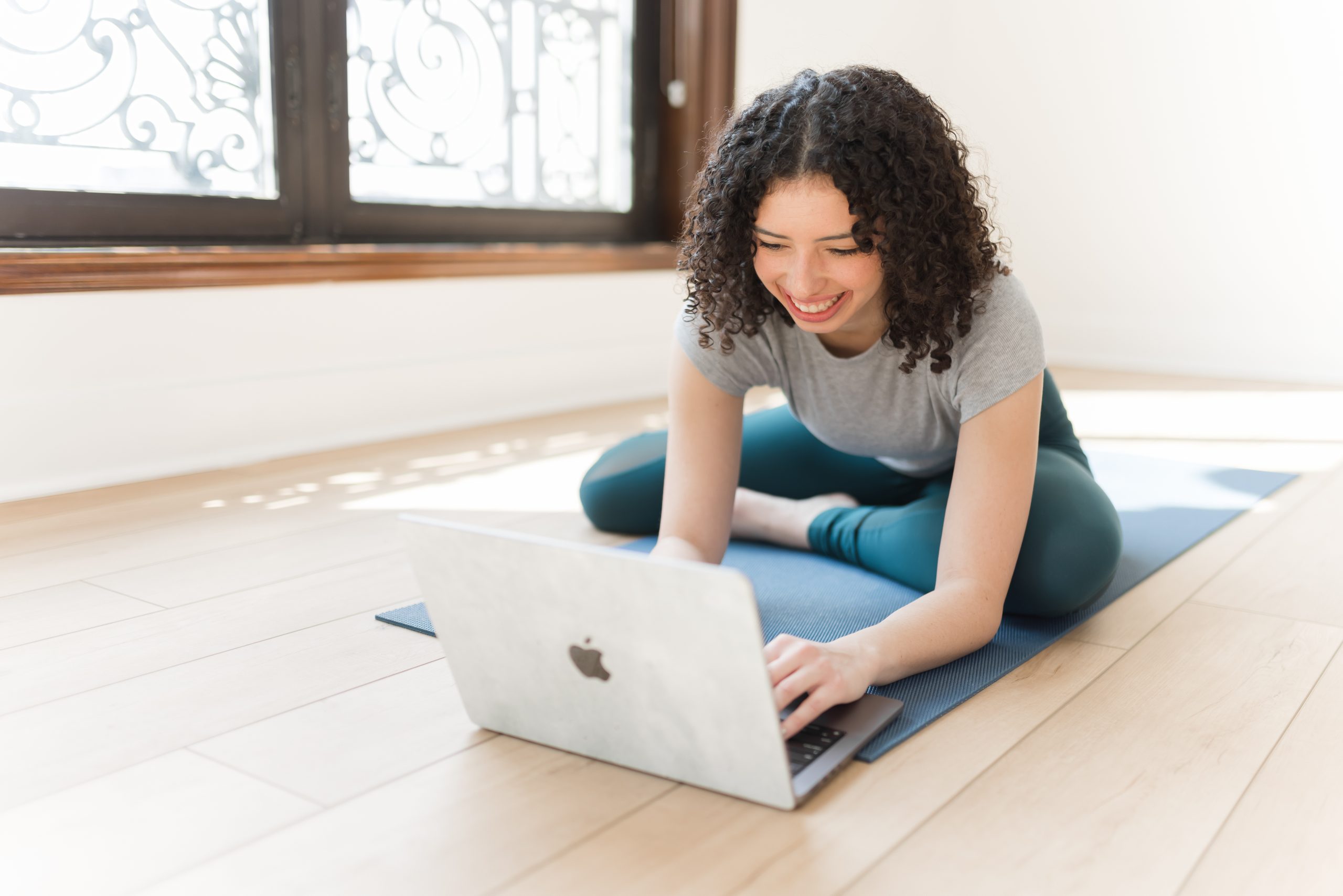 A woman on her laptop sitting on a yoga mat smiling