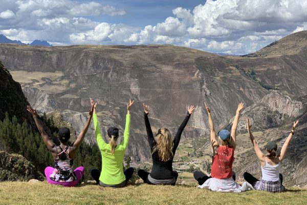 A bunch of yogis on a yoga retreat with their backs to the camera, sitting on a mountainside with their arms up in Urdhva Hastasana