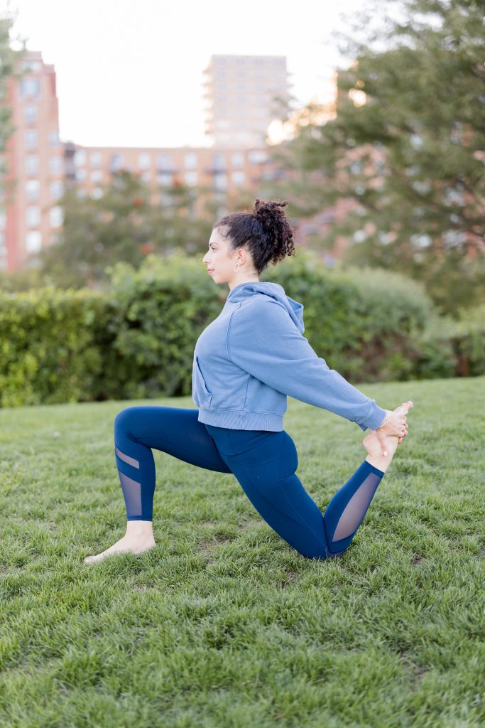 Girl in a blue sweatshirt and blue yoga pants in Anjaneyasana grabbing her foot behind her