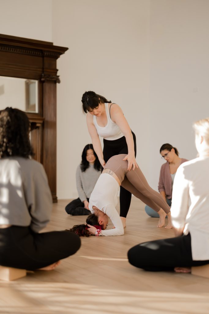 A reliable teacher subbing for another yoga teacher and assisting a student in downward facing dog