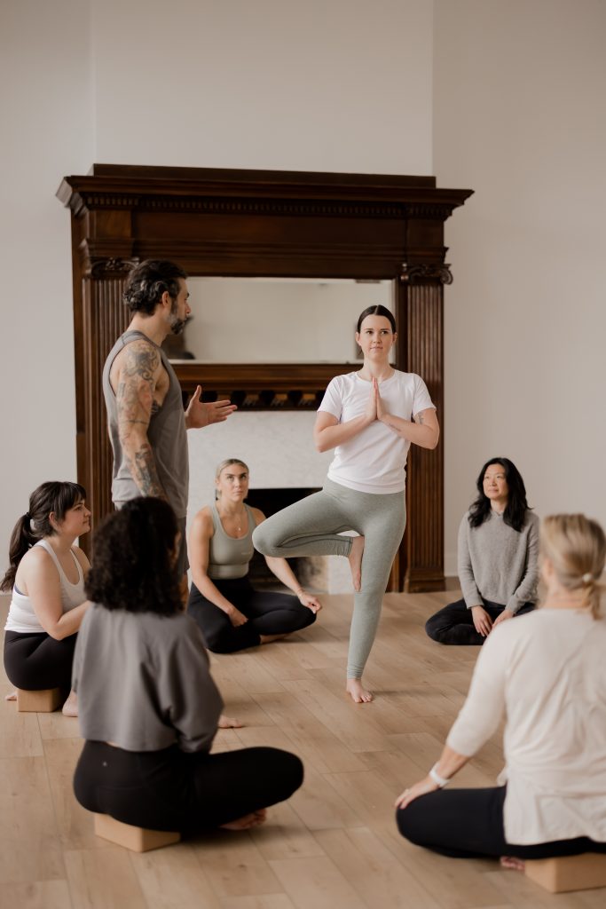 A male yoga teacher continuing to practice his yoga skills by instructing a student to get into tree pose